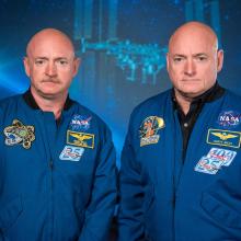 Two adult men who are identical twins stare at the camera. They are both bald, the one on the left has a mustache. They wear blue flight jackets with NASA patches on them. 