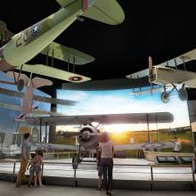 A digital rendering of a museum gallery shows several people standing in front of a display of three vintage biplanes. Behind the airplanes is a huge, floor-to-ceiling projection screen.