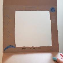  Square piece of cardboard with a square opening cut into the middle of the cardboard. A planet, a moon, and yellow and blue decorations have been hand drawn around the border of the opening. The words, "My Night Sky" are hand written on the bottom of the square opening. 