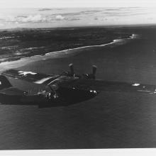 Small dark-colored plane flying over water.