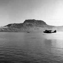 Wide view of an aircraft flying over water in the distance.