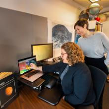 Two researches look at a computer monitor with colors displayed on it. In the background is a topographical map of a planet and globes of different planets. 