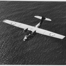 Black and white image of a small aircraft viewed from above flying over water.