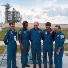 Five men in blue NASA flight suits stand grouped together. A launch pad with a space shuttle stack on it is seen in the distance behind them.