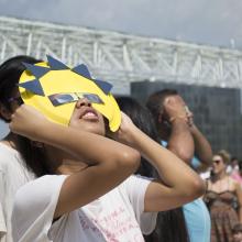 A young woman looks through a pair of eclipse glasses framed by a cutout of the sun.