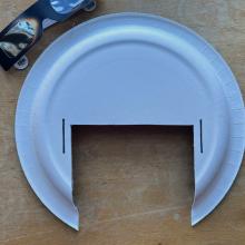 Photo of white paper plate with a section cut out of it. There are a pair of solar eclipse glasses at the top of the photo that you can see partially. 