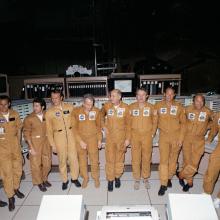 10 middle-aged men wearing yellow-brown jumpsuits stand in Mission Control. Some are looking at the camera and others are looking away.