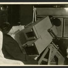 A person sits by the window of an airship with a large camera device pointed at the window.