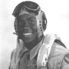 A man poses for a portrait wearing a flight cap and goggles and a flight suit.