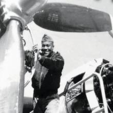 A man in a military uniform faces the camera and smiles as he works on an airplane propellor. 