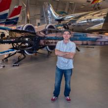 A middle-aged man in a cap, blue short sleeve button up, and jeans, stands with his arm crossed in front of a drone styled to look like a life-size x-wing from Star Wars.