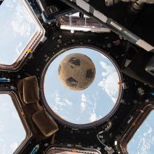 A soccer ball floats inside the International Space Station surrounded by a panoramic array of windows that provide a view of Earth and space.