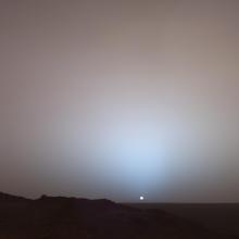 Mars. A landscape under a hazy sky with the sun visible as a small, bright dot near the horizon. The terrain appears to be barren and rocky. The atmosphere is diffusing the sunlight, creating a soft glow around the sun.