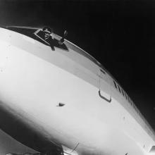 A photo of a man in a pilot's uniform leaning out of the window of an aircraft flight deck as he gives a thumbs up.