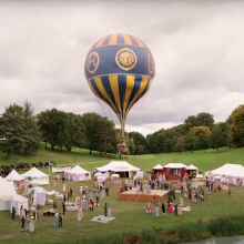 A vibrant hydrogen balloon with intricate blue and gold designs floats above a bustling outdoor event set against a cloudy sky. Below, several white tents are set up in a grassy park, with groups of people mingling and walking around.