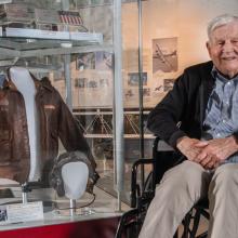 An elderly individual in a wheelchair smiles while seated next to a display of aviation memorabilia, including a leather jacket and a pilot helmet, inside a museum setting.