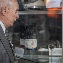 An elderly individual, Bill Anders, looks at a watch on display in a case amongst other space artifacts including gloves and helmets.