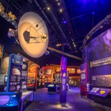 A spacecraft with a large dish and a long boom hanging in a space gallery lit with purple and orange light. 