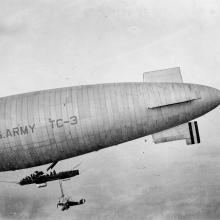 A large airship with US ARMY TC-3 written on the side a small aircraft hangs under the airship.