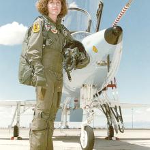 A woman in her mid-20s, wearing a pilot jumpsuit, stands in front of a military aircraft.