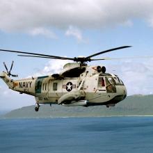 A white and gray helicopter flies over water with the coastline in the background.