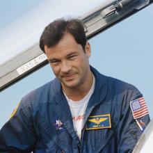 An individual in a blue flight suit with NASA and American flag patches, standing next to part of an aircraft with the sky in the background. 