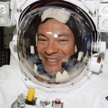 An astronaut wearing a spacesuit and smiling at the camera. The suit features patches of the American flag and another emblem with a star. The background shows equipment and parts of a spacecraft interior, suggesting this image was taken during a space mission.