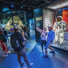 A group of people in the Destination Moon gallery. Two people take a selfie with Neil Armstrong's spacesuit, which is in a case.