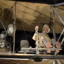 A pilot sits exposed on the lower wing of a vintage aircraft.