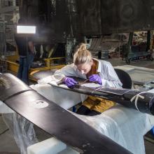 A woman with blonde hair, leans down in intense focus on the propeller of an airplane. The rest of a the conversation hangar fades out around her, you can feel her focus. 