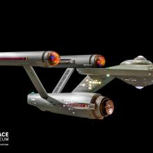 A studio model of the spaceship Enterprise from Star Trek floats against a black background. The ship as a disk shaped center with three horizontal cylinders trailing behind.  