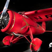 A red monoplane with a propeller sits at a jaunty angle against a black background. 