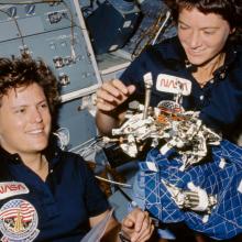 Sally Ride and Kathryn Sullivan, both of whom are white female astronauts, display an object in space.