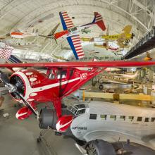 Red sporting monoplane with white trim and clipped wings on display in aviation hangar