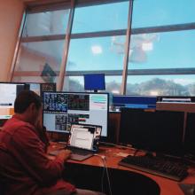 Young man sitting at a desk with computers. Telescope of the Arecibo Observatory can be seen out the window.