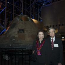 A couple consisting of a white female (left) and male (right) post in front of the conic-shaped metal Apollo Command Capsule.