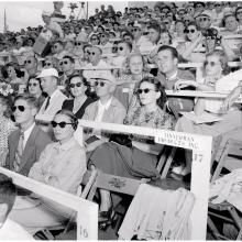 Spectators in a grandstand look to their left. Many wear sunglasses.