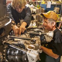 Two white, male Museum specialists work together to perform restoration work on an engine of a World War II bomber aircraft. One of the men is holding his hands below the visible section of the engine while the other man has his fingers above the visible engine section.