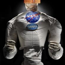 Frontal view of a body of a robot with arms, chest with a NASA logo, and an orange face cover.