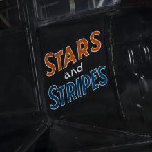 The words Stars and Stripes are painted orange and blue on the side of an airplane. 		