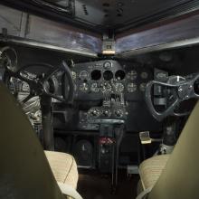 View of the cockpit on the plane. The image was taken behind the seats of the cockpit. The image highlights the instrument panel of the plane. 