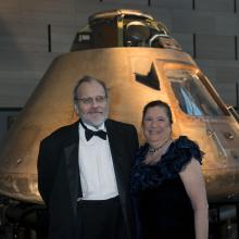 Two people, a white male and female, pose in front of the Apollo Command Module, a conic-shaped metal spacecraft.