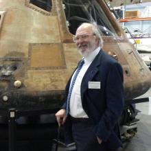 Board member Bruce R. McCaw, an older white male with a beard, poses informally in front of the Command Module Columbia