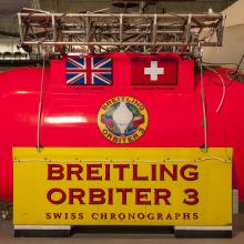 Bright red and yellow metal structure that is the gondola used to carry people and equipment during a balloon ride. It has the inscription, Breitling Orbiter 3. Attached to the metal structure are silver tanks.