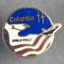 A small gold pin decorate with red, white, and blue. It depicts an artistic interpretation of an eagle flying across an American flag and a space shuttle flying in space.