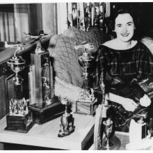 Black and white image of a woman sitting by a desk with various trophies.