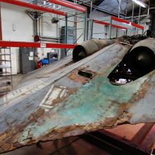 Aft, diagonal view of a gray World War II-era aircraft which reveals green paint under where a fairing section was located prior to its treatment.