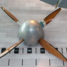 Large, four-blade wooden fan used for a wind tunnel that is hanging in the museum. The center is a silver-colored metal base.