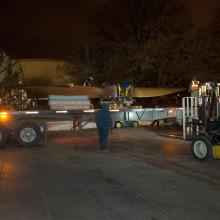 Outside of the Museum, a wind tunnel fan is transported by a truck with a large flatbed into the Museum.