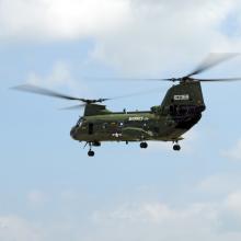 Side view of green military helicopter in flight. The helicopter has two separate propellors, both of which have three blades.